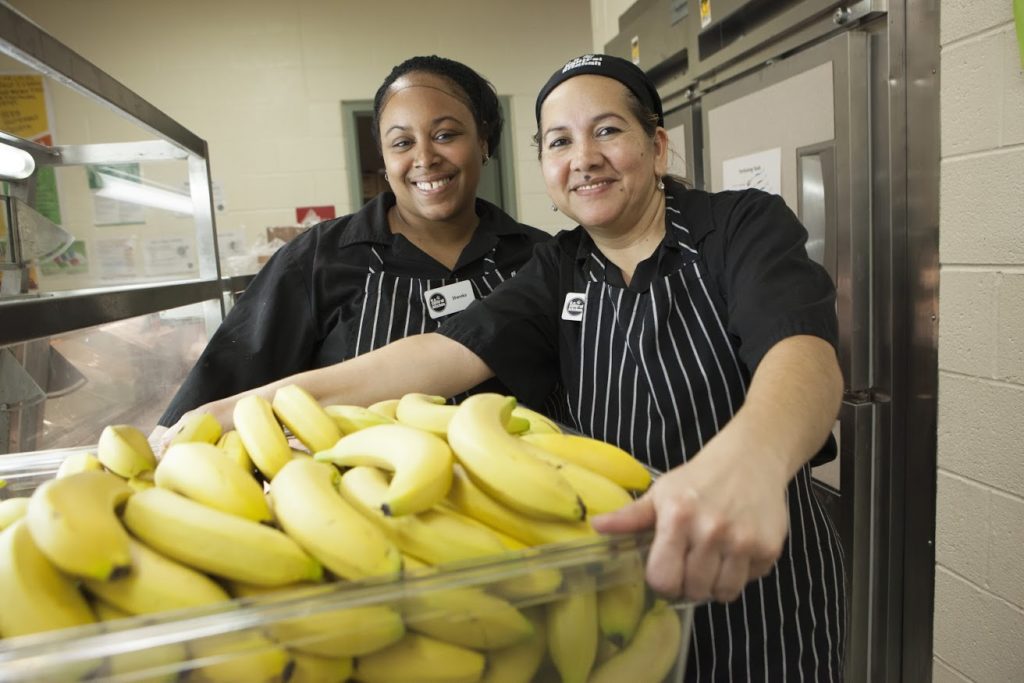 DC Central Kitchen photo of two staff with bright yellow bananas in bin at forefront of photo, quarter 3 2017 poverty and health beneficiary